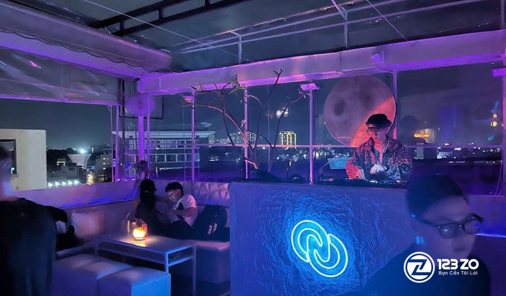 Quán rooftop tại Quận Phú nhuận với ánh sáng tím, khu vực ghế sofa và một DJ đang chơi nhạc ở quầy, có tầm nhìn ra thành phố đêm.